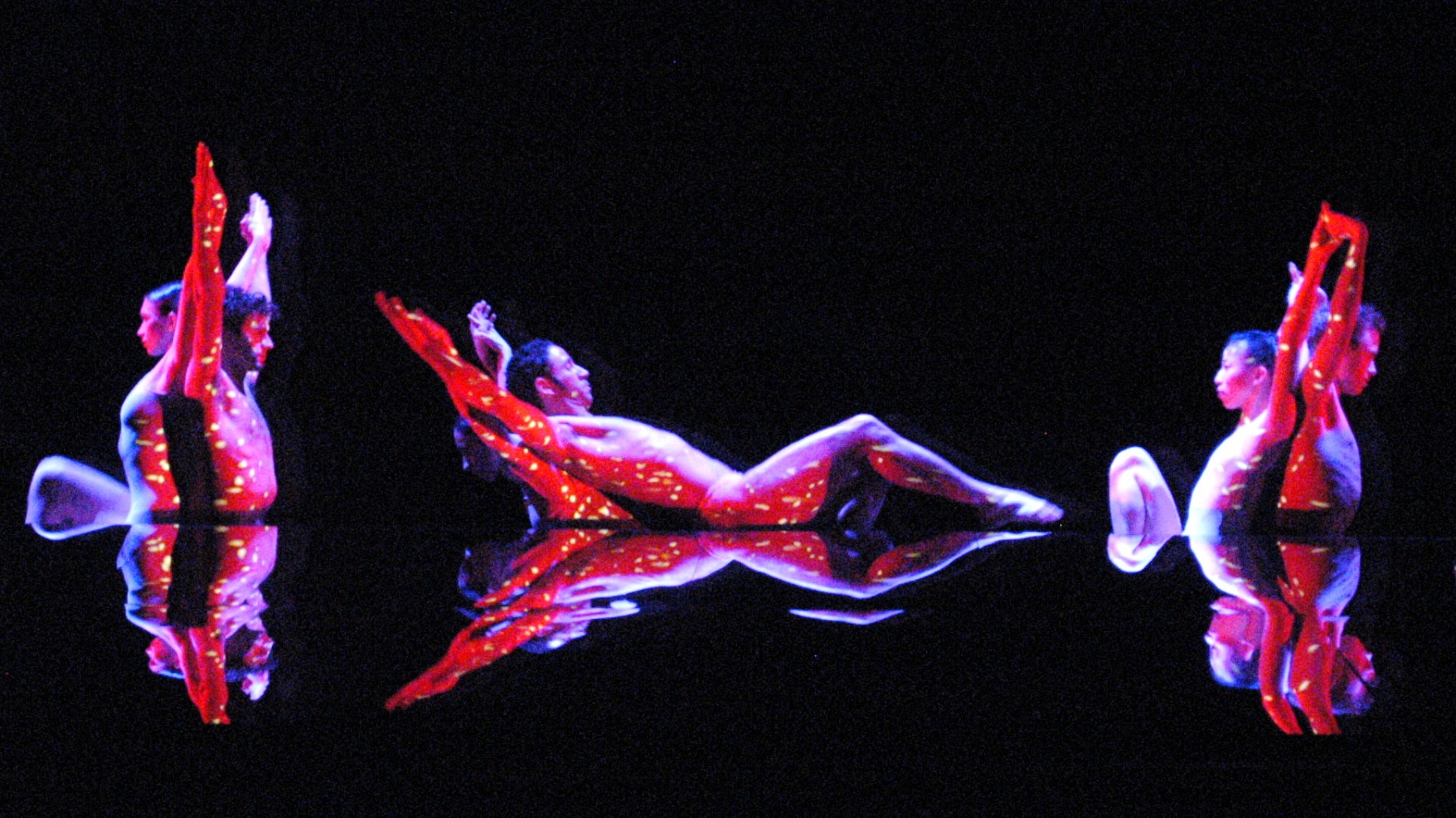 Red and yellow light is projected on dancers' skins as they stand back to back with their arms extended